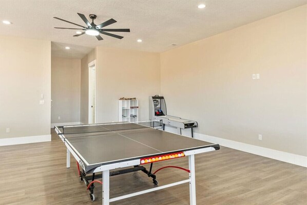 Una mesa de ping pong en una habitación con suelo de madera, un ventilador de techo y un estante con objetos.