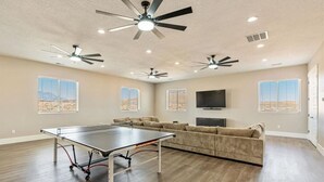 Amplia sala de estar con mesa de ping pong, varios ventiladores de techo, un televisor de pantalla plana y ventanas grandes con vistas al desierto.