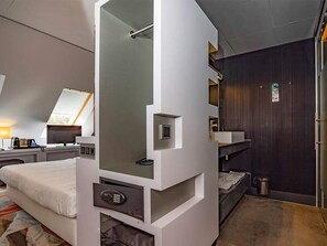 Experience a unique blend of comfort and functionality in this stylish hotel room, featuring a large bed with a mirror and control panel, a cozy seating area, and ample storage space.