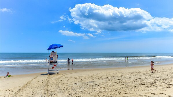 Una playa con un puesto de salvavidas, una sombrilla azul y gente caminando por la orilla.