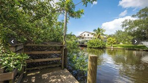 A wooden dock extending over a calm water body, surrounded by lush greenery and a house in the background. - Waterfront Key West Style Home (Ellenton)