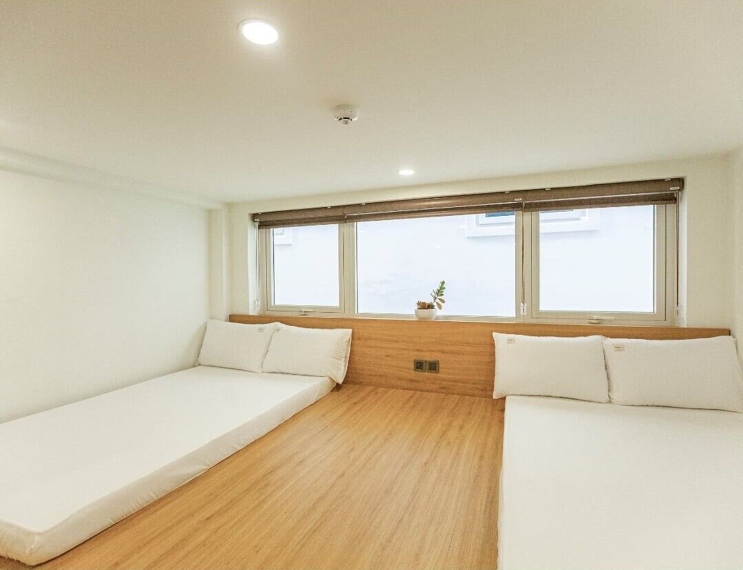 A room with two single beds, a wooden floor, a window with blinds, and a small plant on the windowsill.