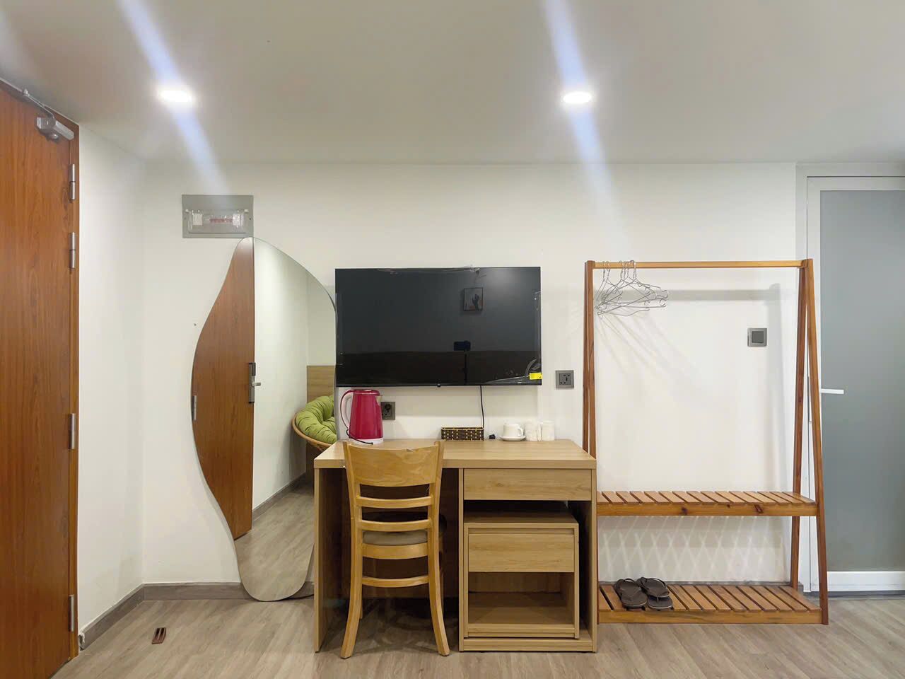 A compact room with a wooden desk, a chair, a TV mounted on the wall, a hanging rack, and a door with a mirror.