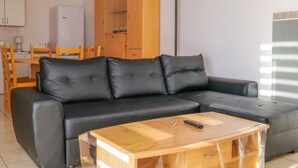 Un salon moderne avec un canapé en cuir noir, une table basse en bois et un coin repas comprenant des chaises en bois et un réfrigérateur blanc.