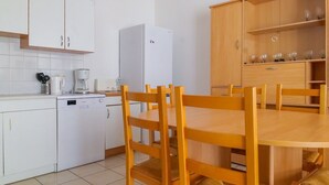 Une cuisine de taille réduite avec des meubles blancs, un réfrigérateur blanc, une machine à café et une table à manger avec des chaises en bois.