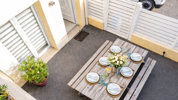 Une table en bois, dressée pour six personnes, avec des assiettes, des couverts et un vase de fleurs, le tout entouré d’un mur jaune et d’une porte blanche.