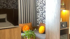 Une chambre d’hôtel compacte comprenant un lit, un coin salon avec des coussins, une petite table avec une plante et une armoire avec un miroir.