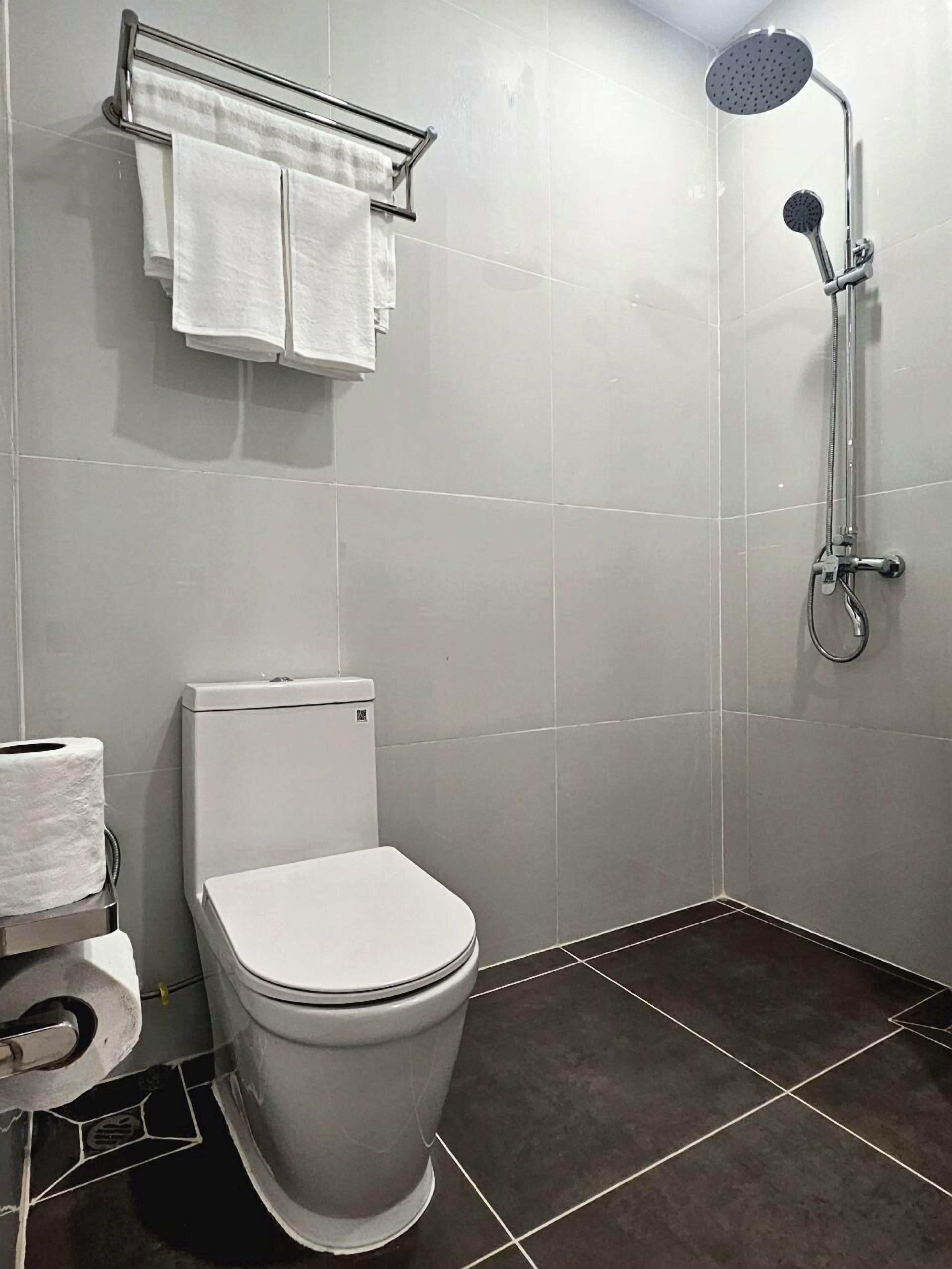 Um banheiro moderno com vaso sanitário branco, chuveiro com chuveiro manual e porta-toalhas com duas toalhas brancas.