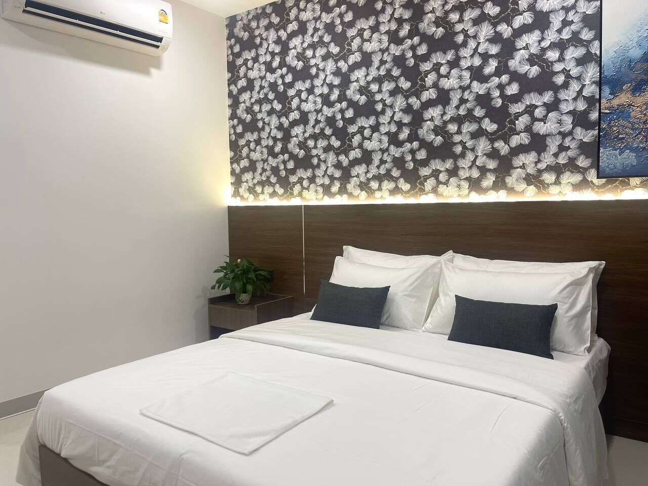 Habitación de hotel con una cama grande, una mesita de noche con una planta, un aire acondicionado de pared y un papel tapiz con motivos florales sobre la cama.