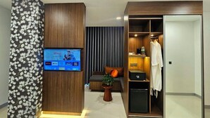 Une chambre d’hôtel moderne dotée d’un téléviseur à écran plat fixé sur un panneau en bois, d’un coin salon avec un canapé et une petite table, ainsi que d’un dressing avec un miroir et une robe accrochée.
