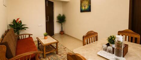 A dining area with a table, chairs, a small rug, a potted plant, and a painting on the wall.