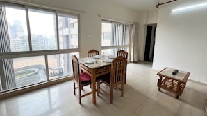Uma área de refeições com uma mesa posta para quatro pessoas, grandes janelas com persianas e vista para prédios e uma rua.