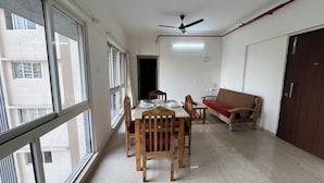 Uma sala de jantar com mesa, cadeiras e um sofá. Há um ventilador de teto e uma porta ao fundo.