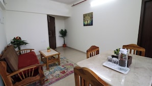 Uma sala de estar com sofá, cadeiras, uma mesa pequena e uma mesa de jantar. Há plantas em vasos e um quadro na parede.