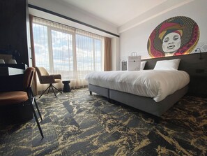 Relax in a spacious room with a large bed, a comfortable chair, and a desk, featuring a window with curtains and a beautiful painting.