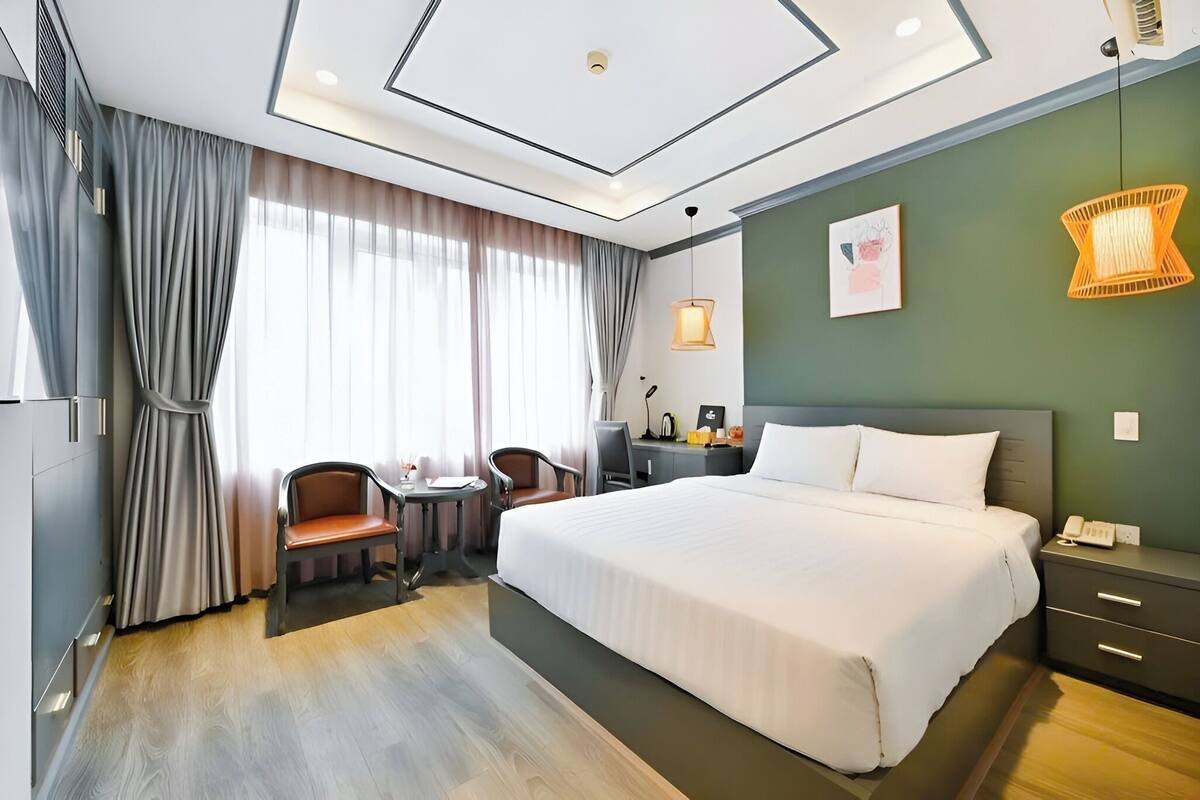 A hotel room with a large bed, a desk with a chair, a small table, a telephone, and a wall-mounted lamp.
