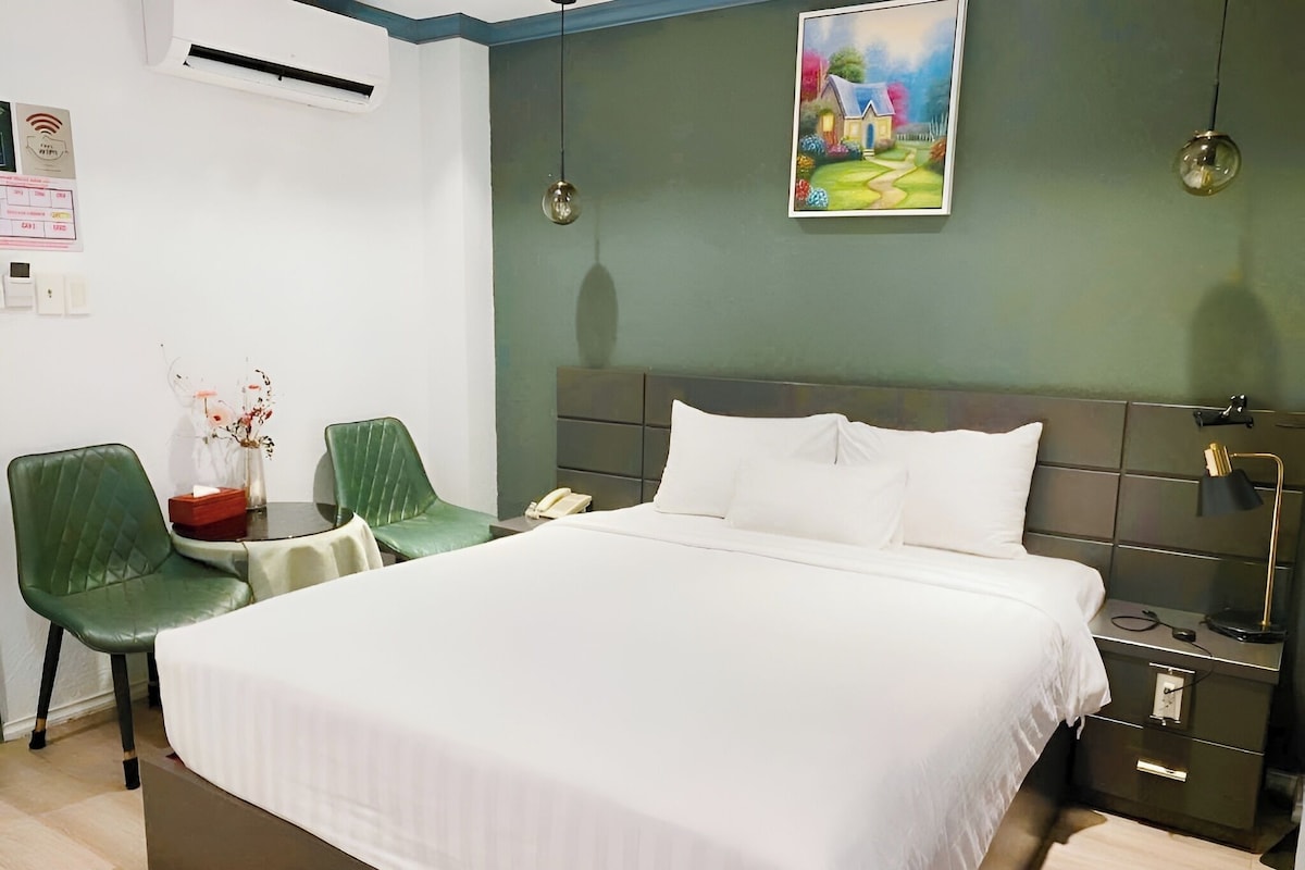 Habitación de hotel con cama, dos sillas, mesita de noche, lámpara, un cuadro y unidad de aire acondicionado.