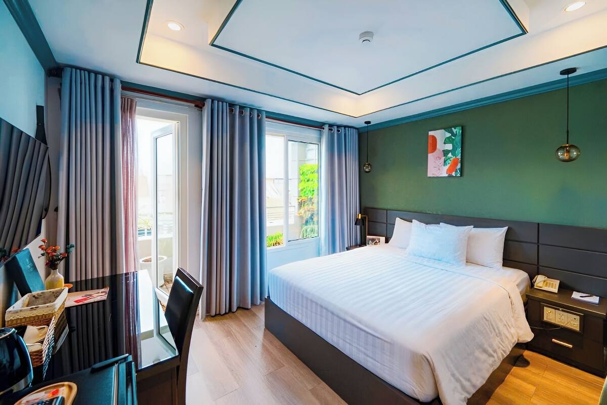 A hotel room with a bed, bedside table, desk, chair, and a view of greenery outside.