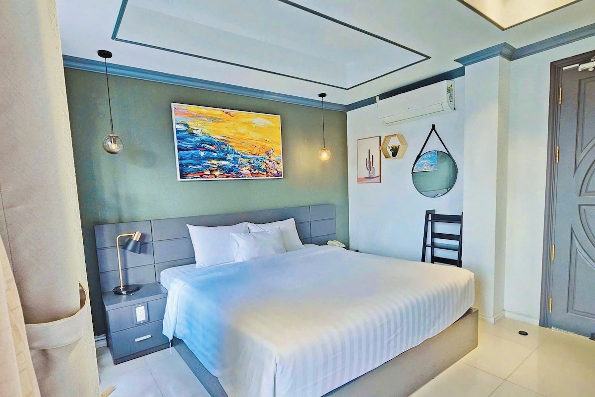 Un dormitorio con cama, mesitas de noche, un cuadro, un espejo y una puerta.
