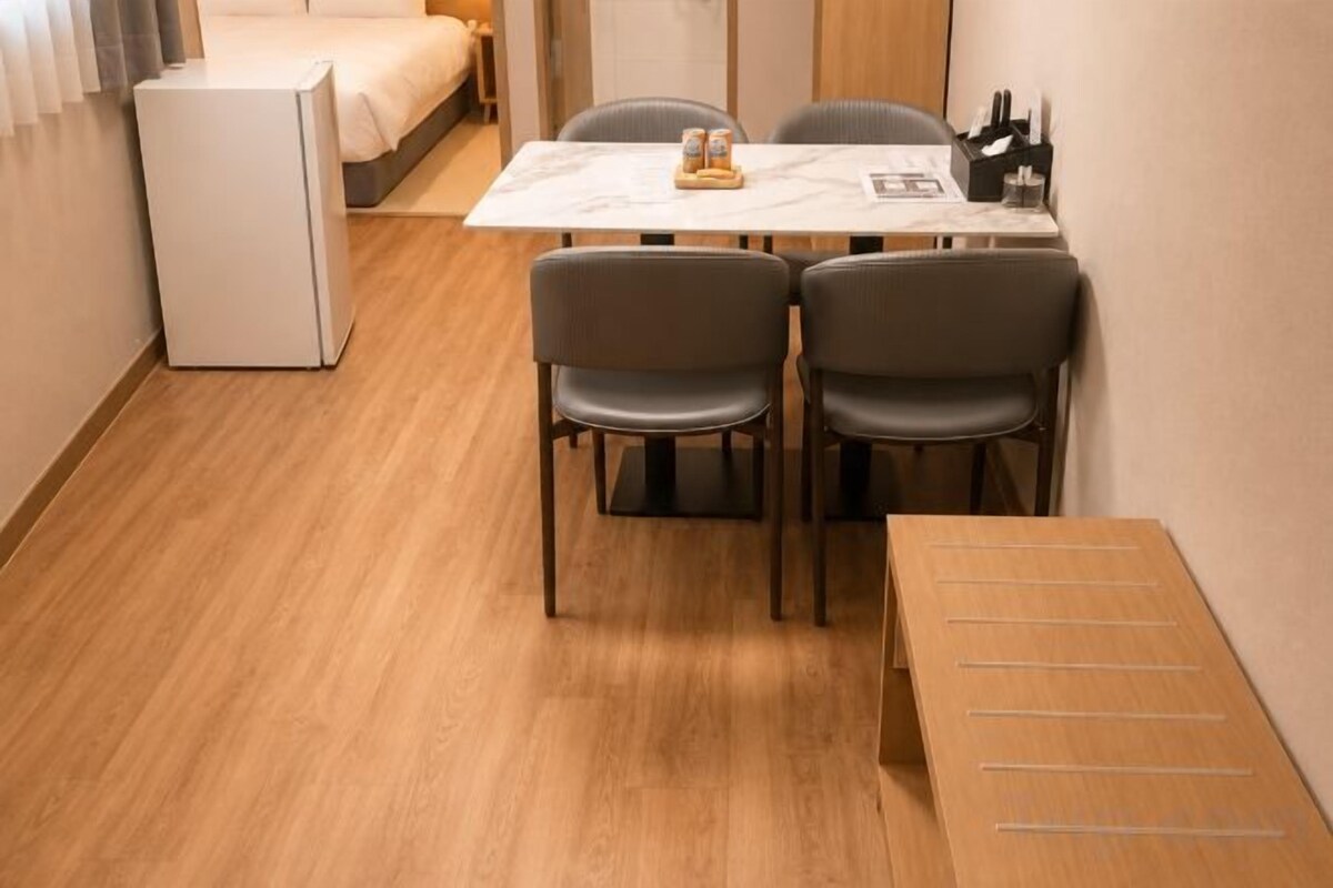 Una habitación compacta con cama, un refrigerador pequeño, una mesa de comedor con sillas y un banco de madera.
