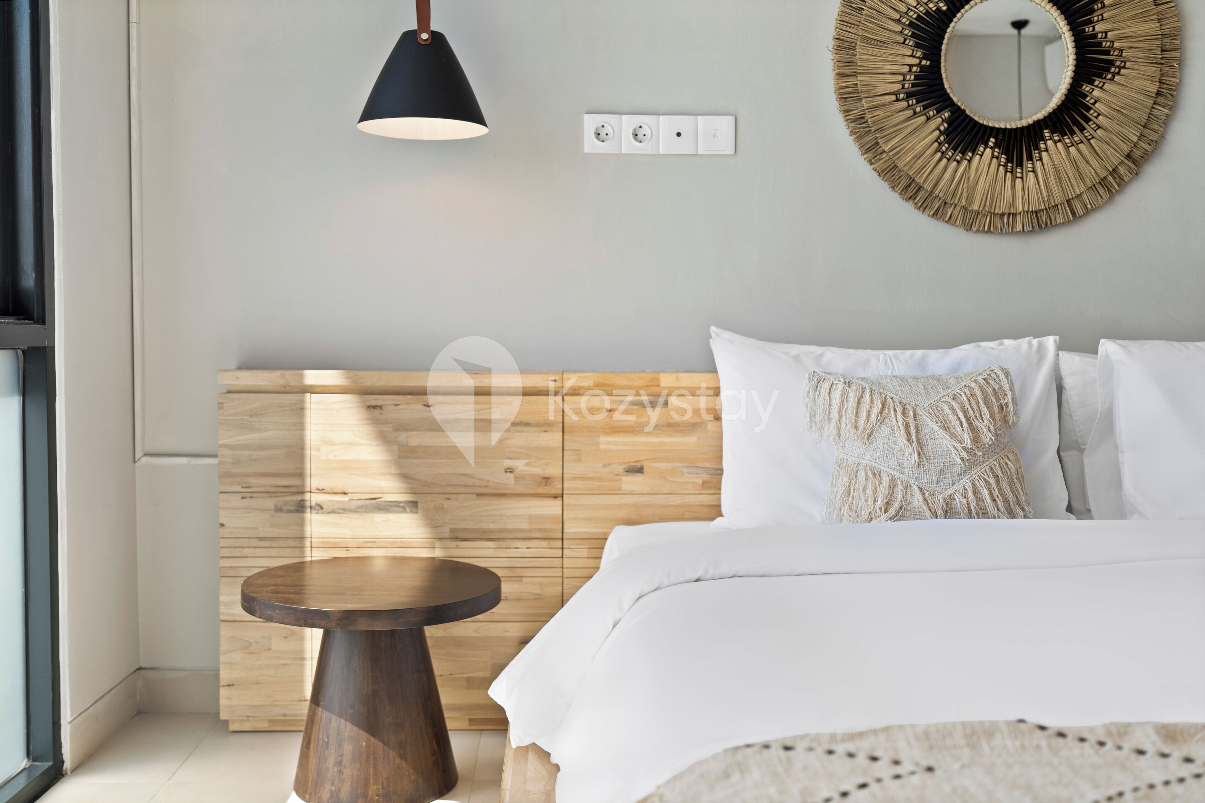 Een moderne slaapkamer met een houten hoofdbord, een rond houten nachtkastje, een ronde spiegel en een hanglamp.