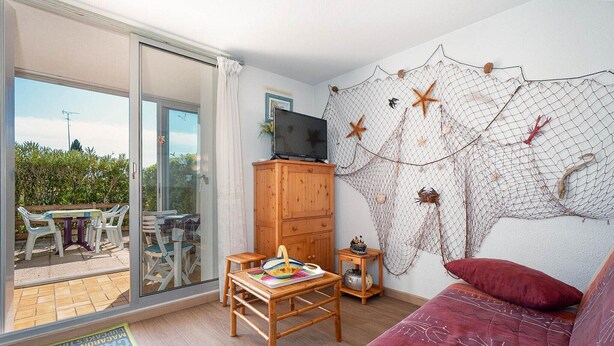 Ein Zimmer mit einem Holzkabinett, einem Fernseher darauf, einem roten Sofa, einem kleinen Tisch mit einem Buch und einer Netzdeko an der Wand.