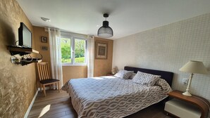 A bedroom with a bed, a chair, a TV mounted on the wall, a window with curtains, a bedside table with a lamp, and a framed picture on the wall.