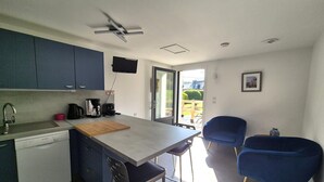 A modern kitchen with a dishwasher, a coffee maker, and a television mounted on the wall. There are two blue chairs and a dining table.