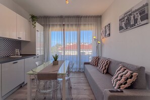 Un soggiorno moderno con angolo pranzo, divano e vista su un edificio filtrata da tende leggere.
