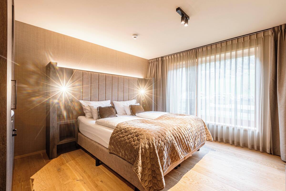 Un dormitorio moderno con una cama grande, cabecero de madera y vistas a la ciudad a través de cortinas translúcidas.