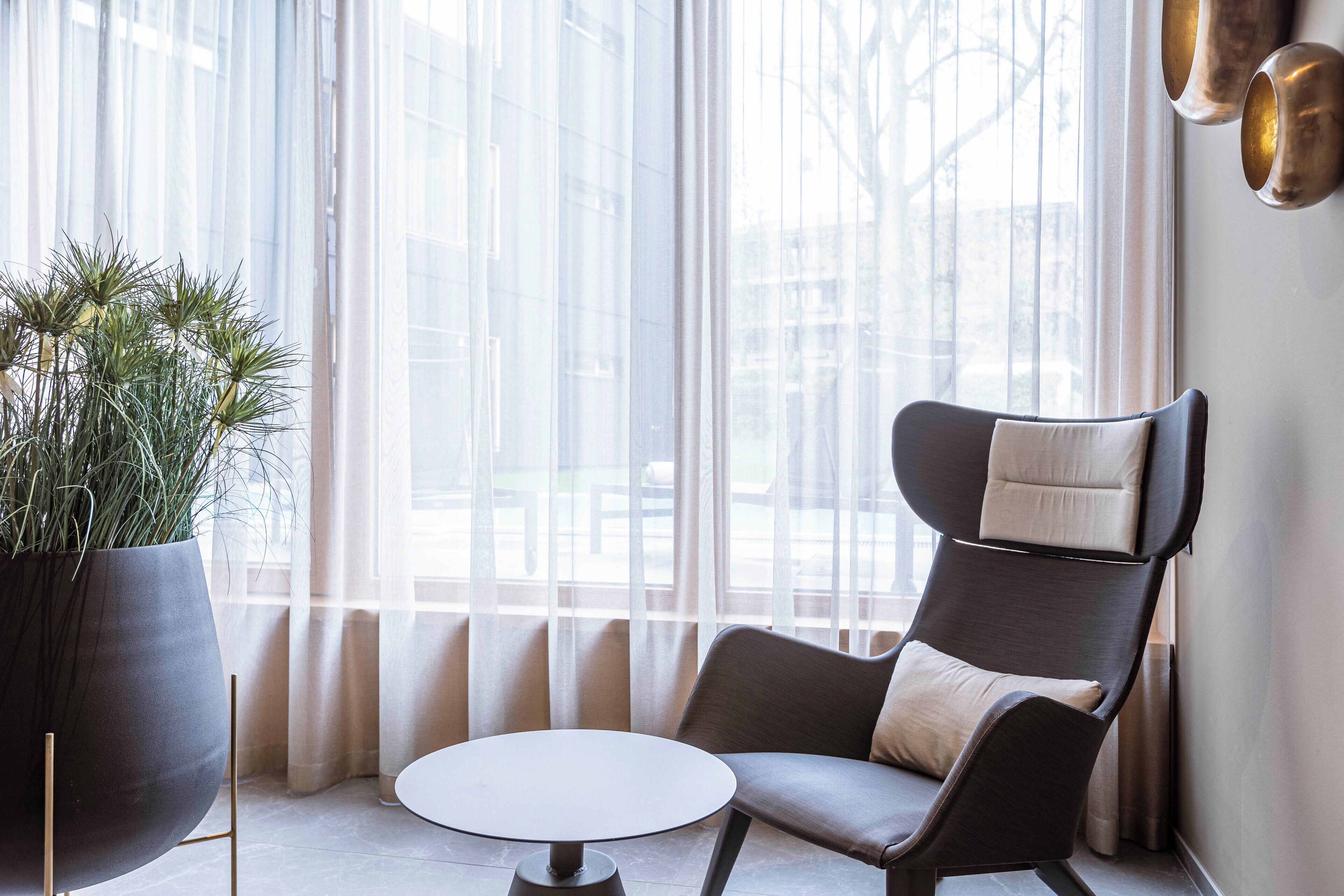 A modern room with a large window, a potted plant, a dark armchair, and a round table.
