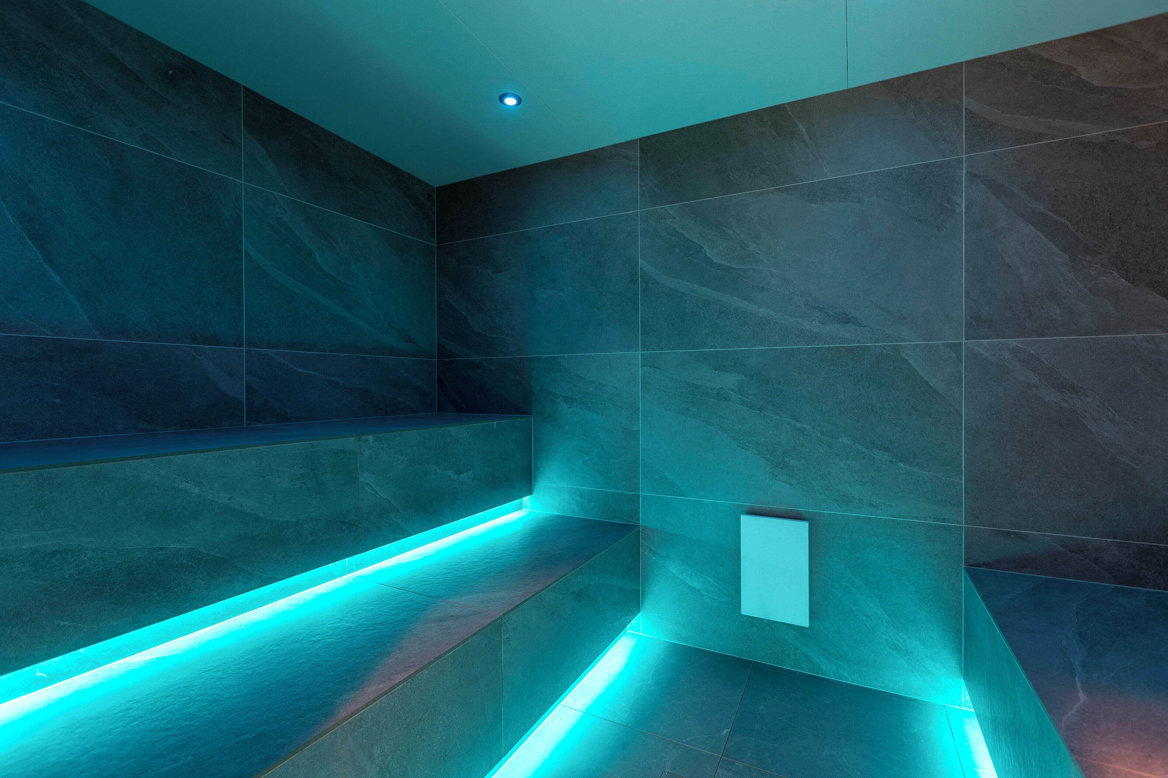 Un baño moderno con baldosas oscuras y una franja de luz LED azul a lo largo del suelo.