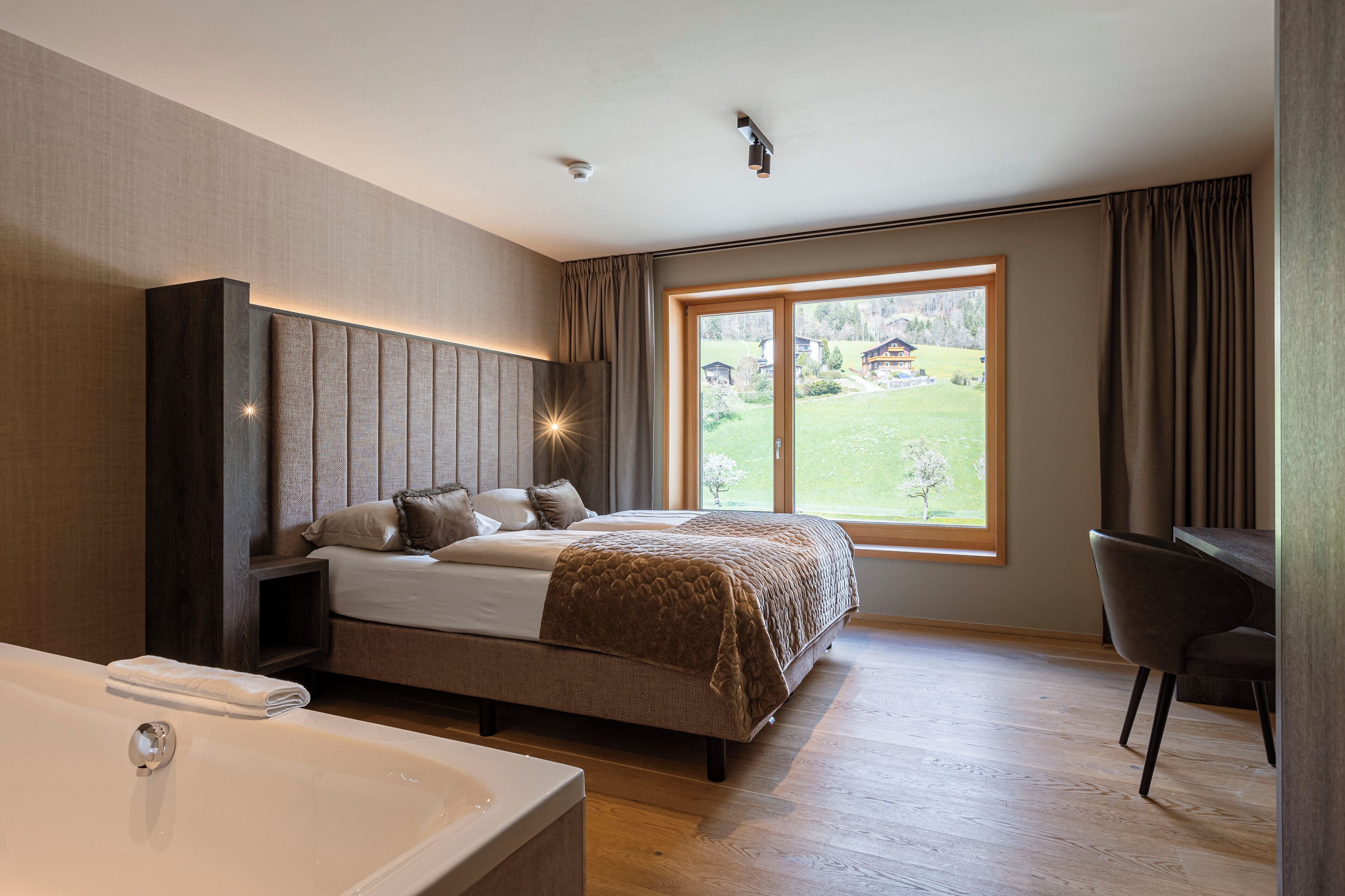 Un dormitorio moderno con una cama grande, un escritorio y vistas a un paisaje verde a través de un ventanal.