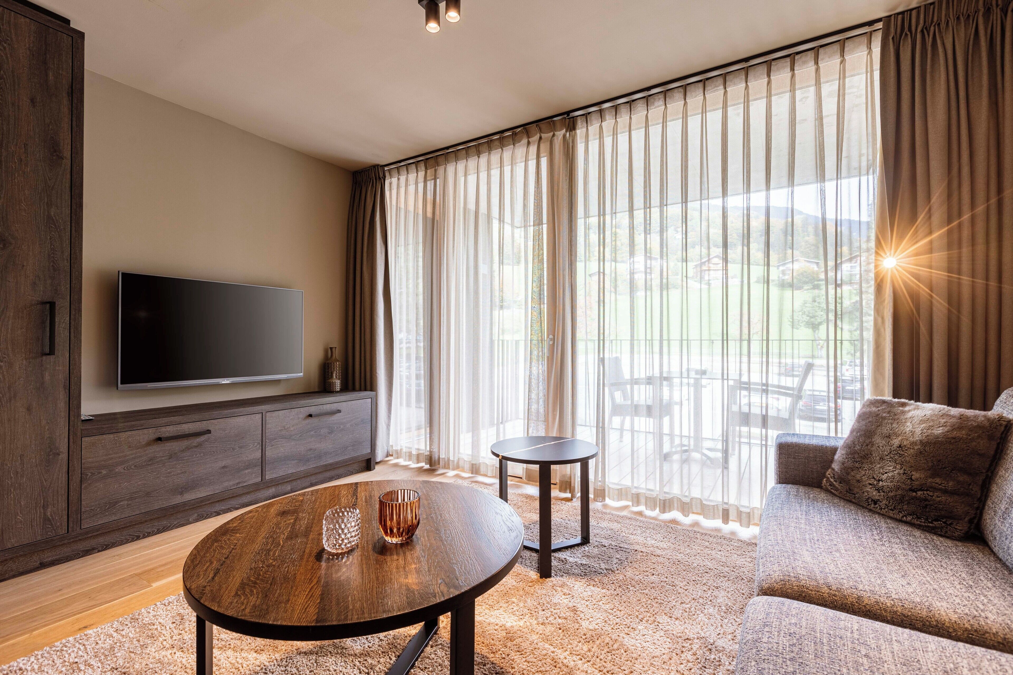 A modern living room with a flat-screen TV, a round wooden coffee table, a sofa, and a view of a garden through sheer curtains.