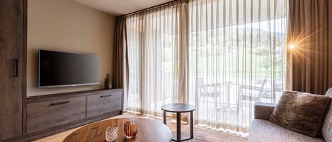 A modern living room with a flat-screen TV, a round wooden coffee table, a sofa, and a view of a garden through sheer curtains.