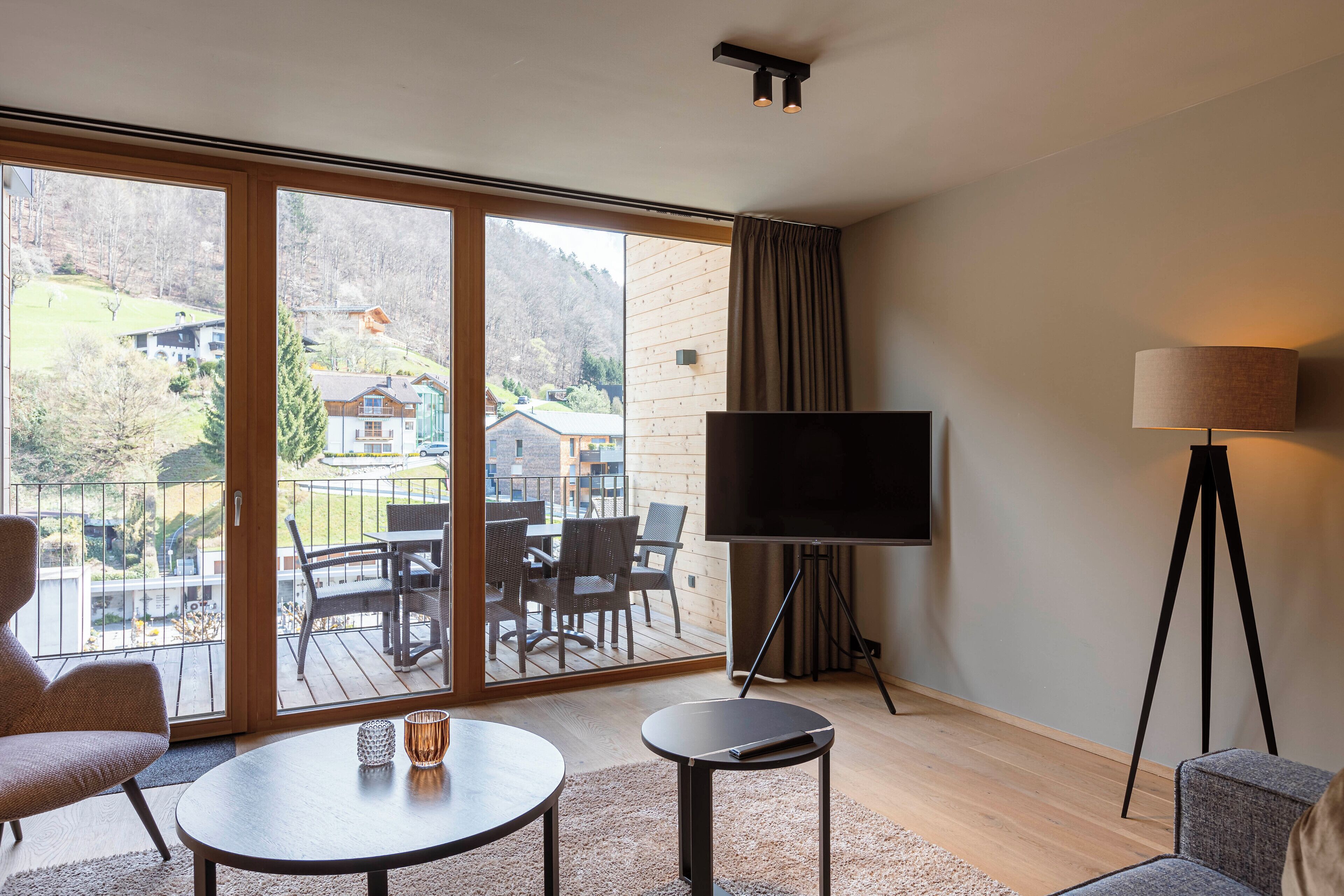 A modern hotel room with a large sliding glass door leading to a balcony, a flat-screen TV, a small round table, and a lamp.