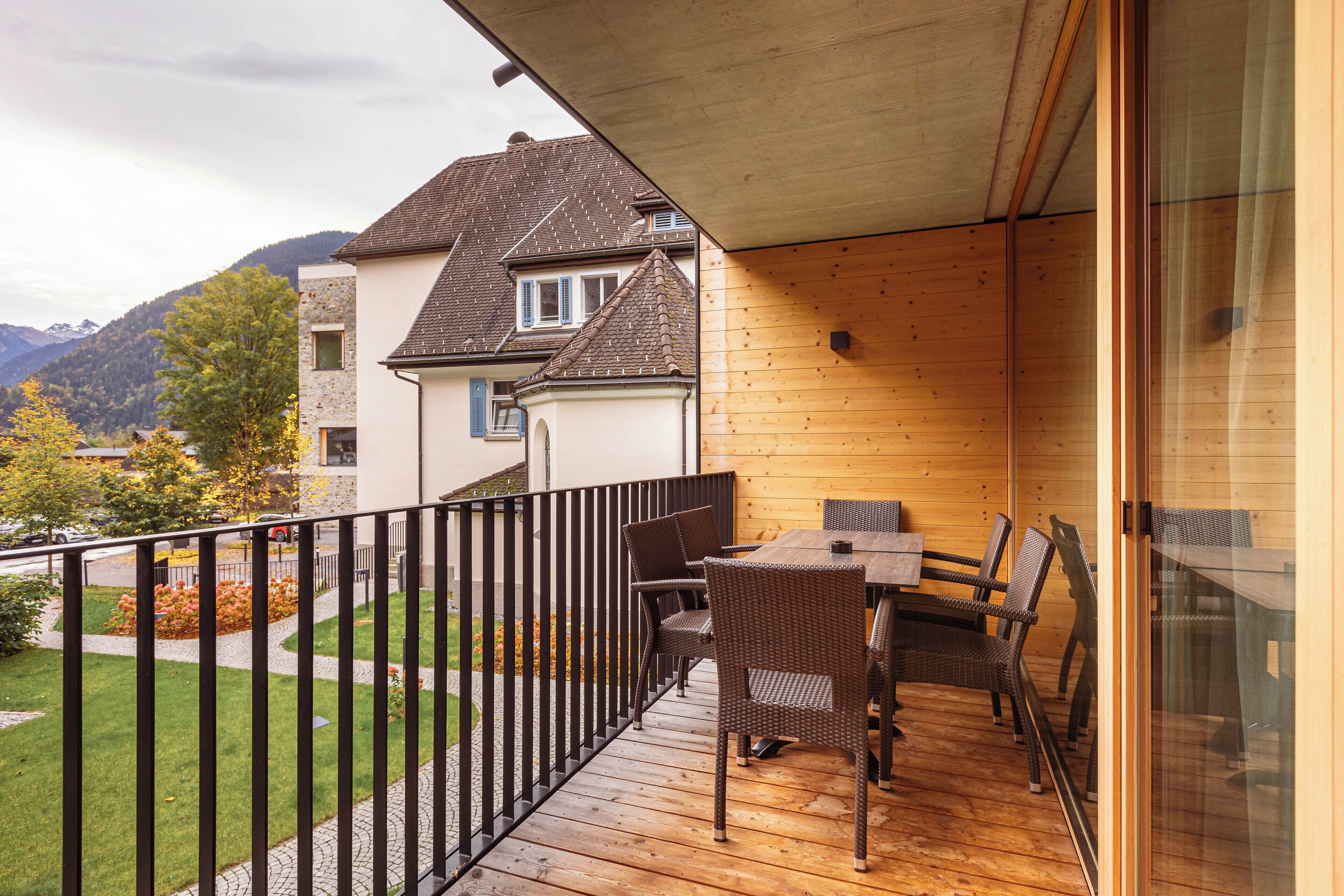 A balcony with wicker furniture, a wooden deck, and a view of a house and mountains.