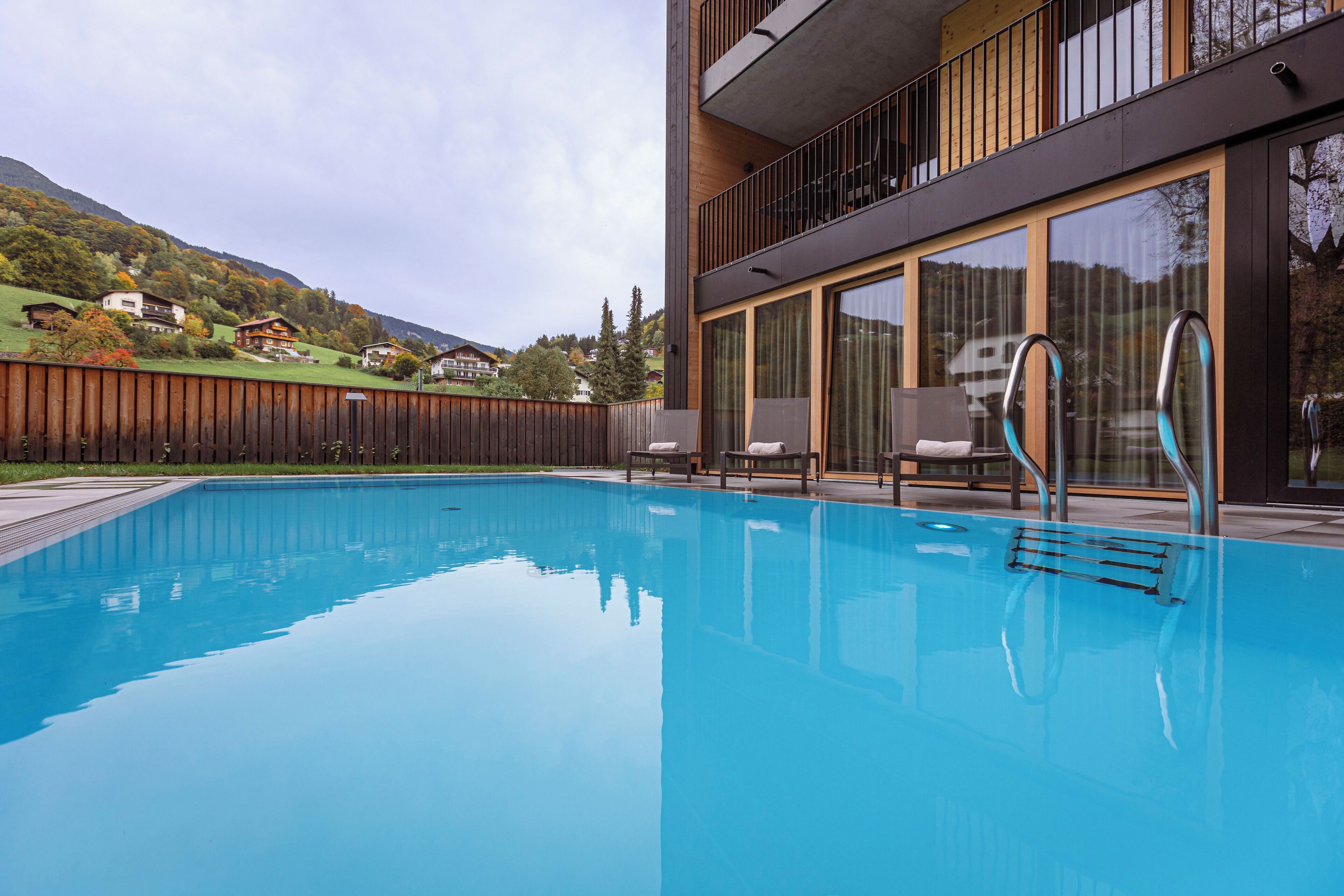 A modern pool area with clear blue water, a wooden deck, and a view of surrounding houses and hills.