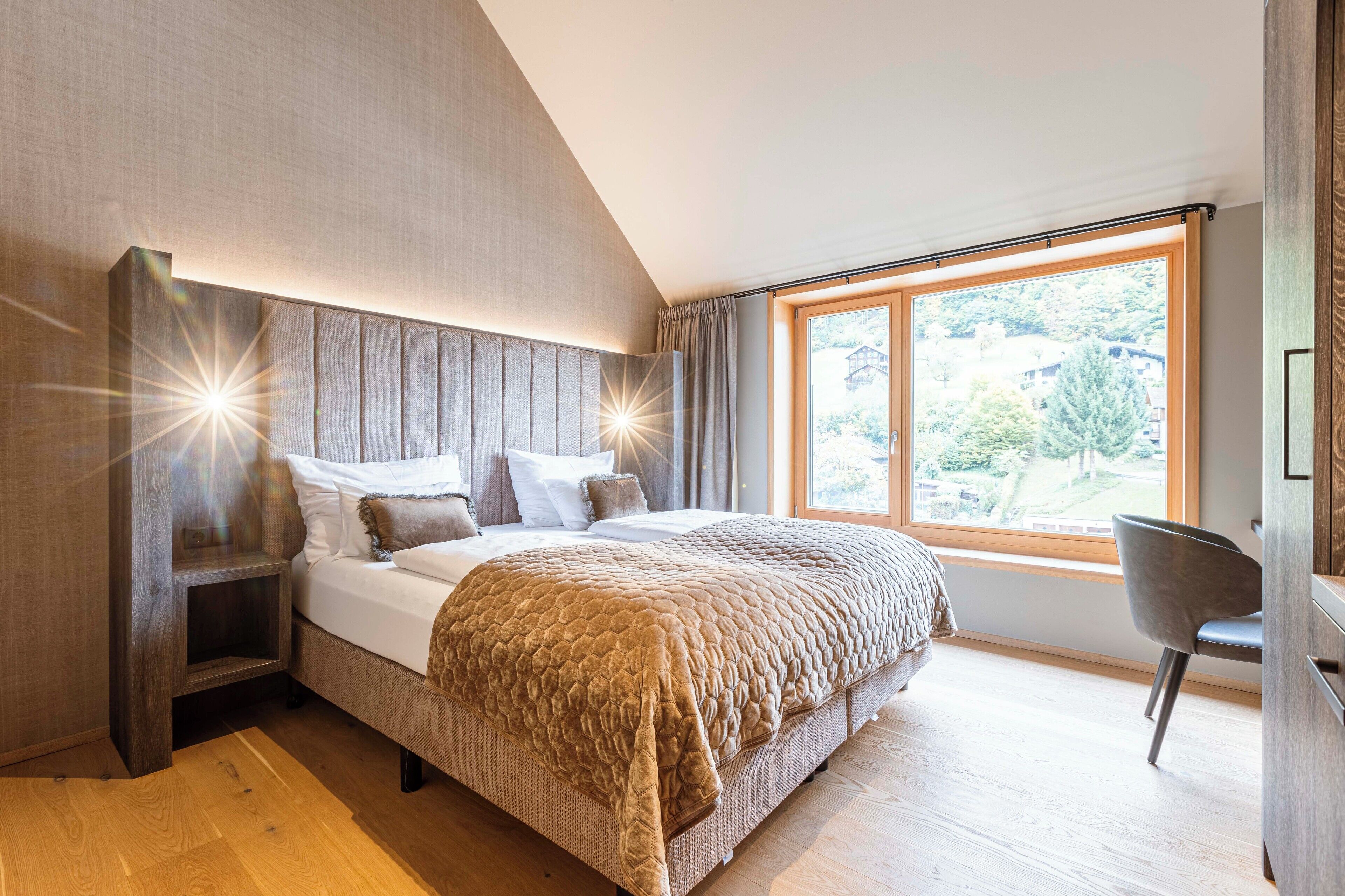 A modern bedroom with a large bed, a nightstand, a chair, and a large window offering a view of greenery.