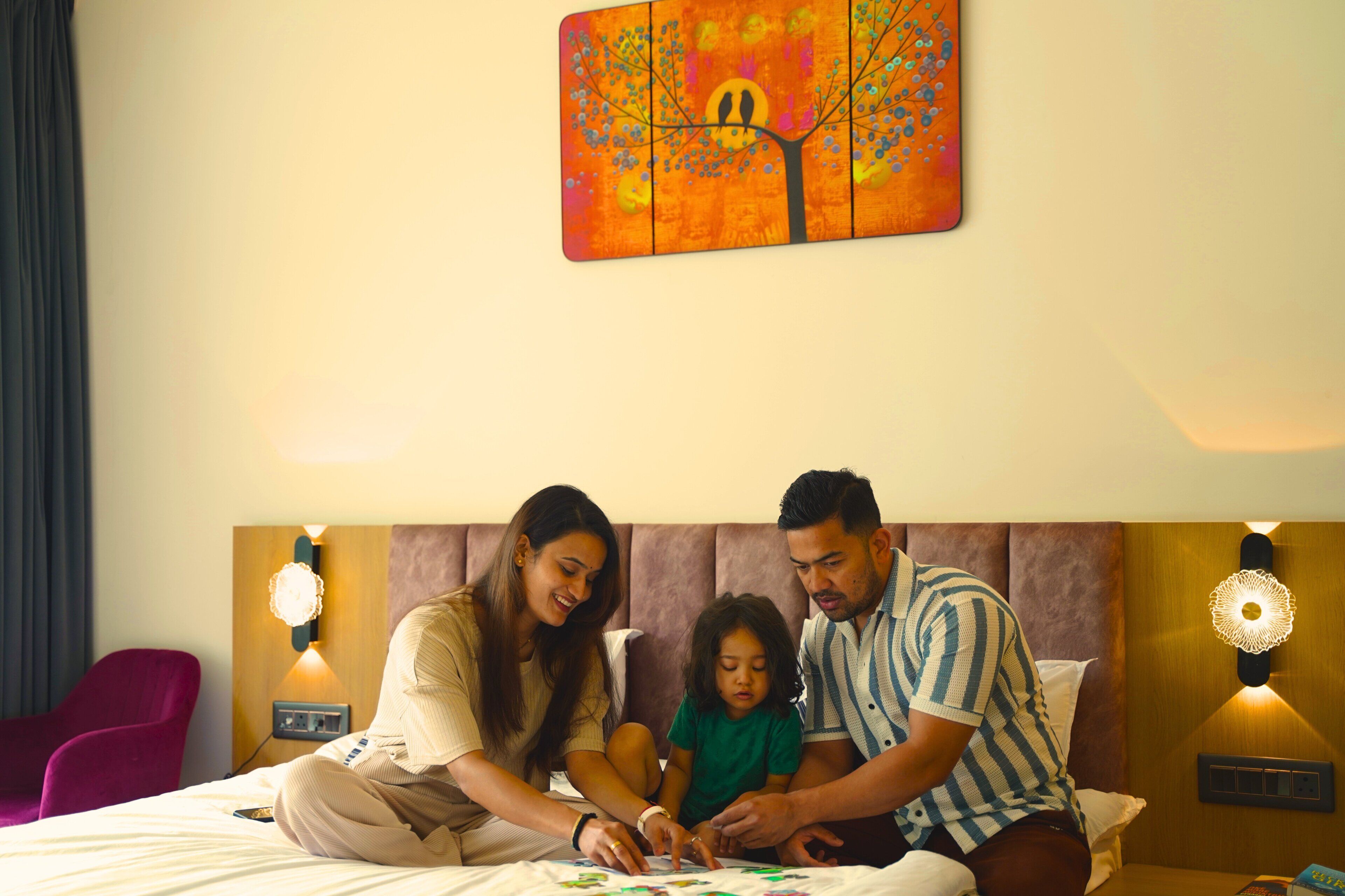 Una familia en una cama, con un cuadro encima, dos apliques de pared y una silla morada.