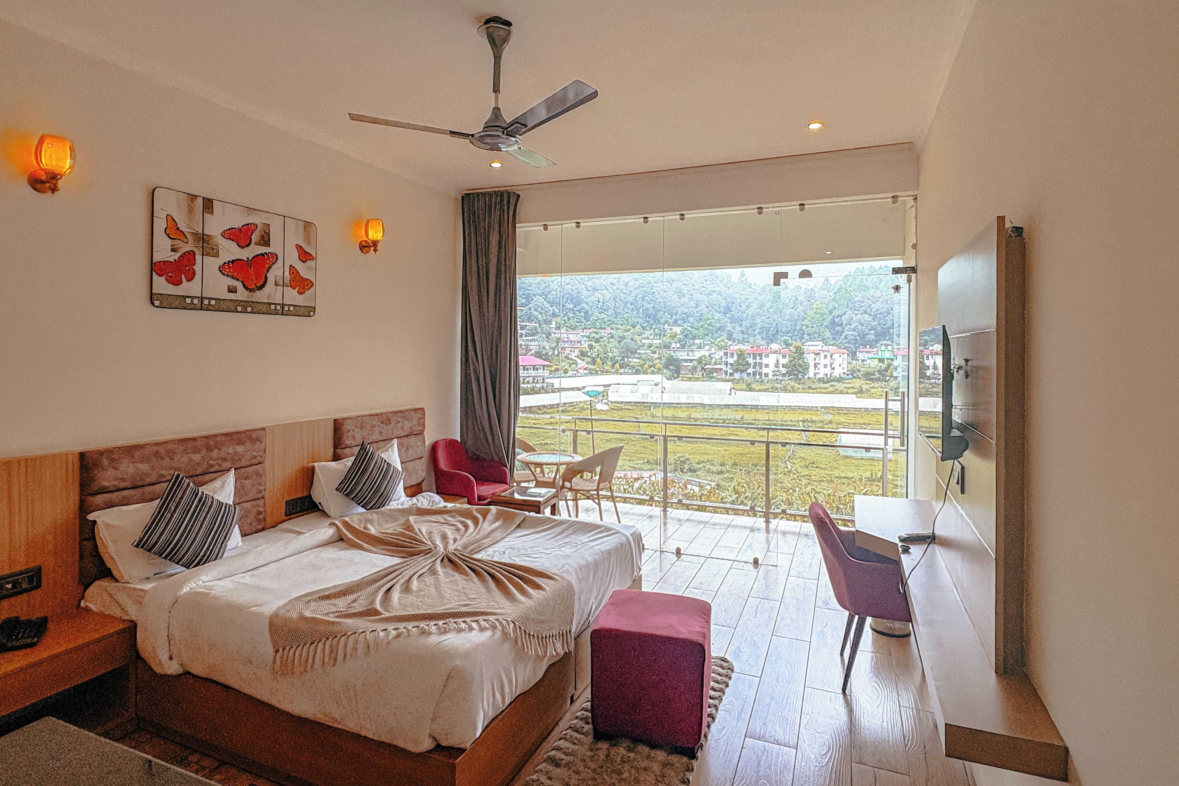 A hotel room with a bed, a desk, a chair, a balcony with a view, and a ceiling fan.