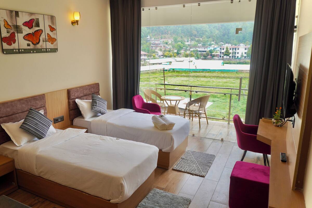 A hotel room with two beds, a balcony with a view of a town, and a dining area with chairs.