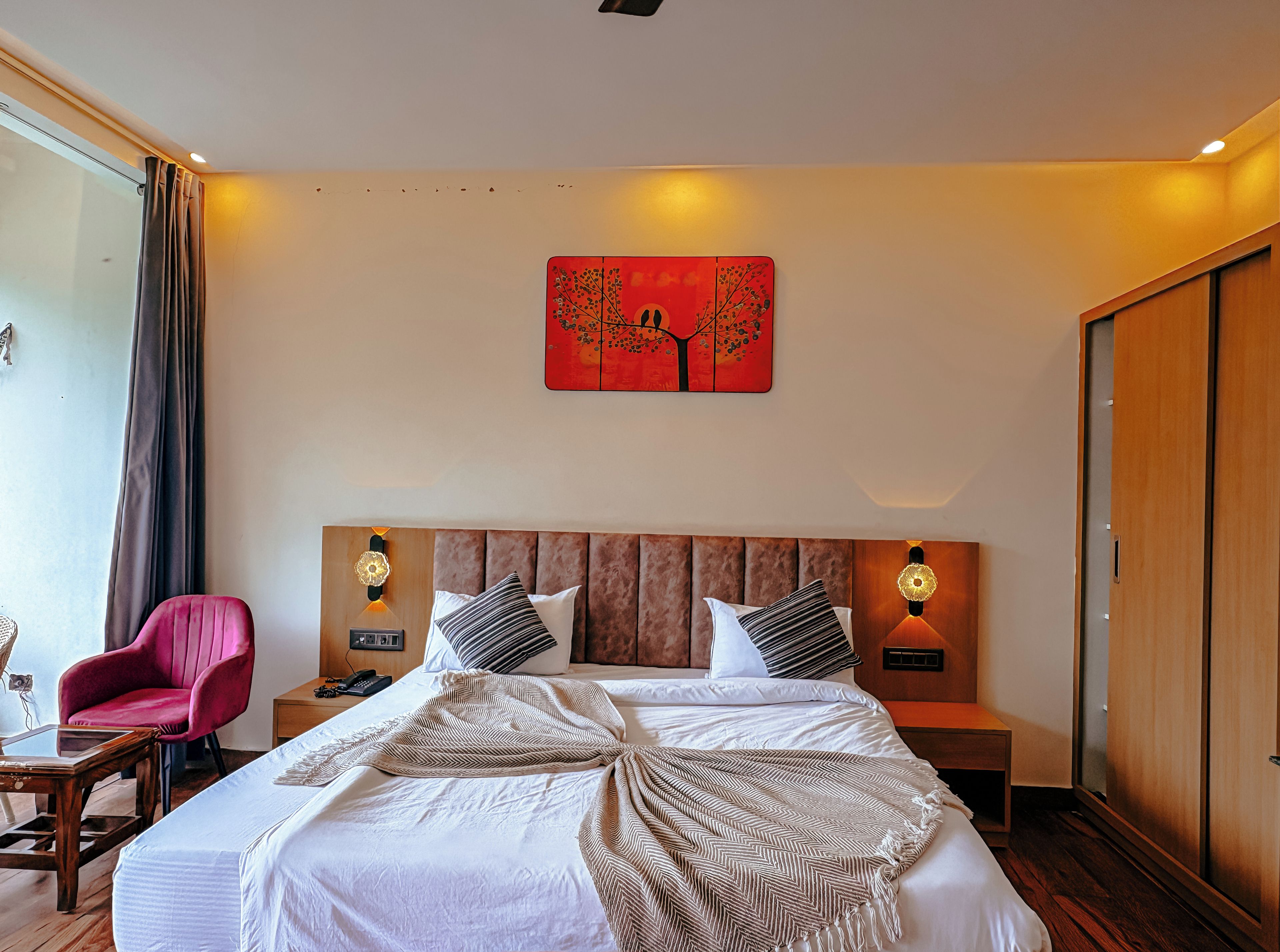 A hotel room with a large bed, two bedside tables, a red artwork on the wall, a pink chair, and a wooden wardrobe.