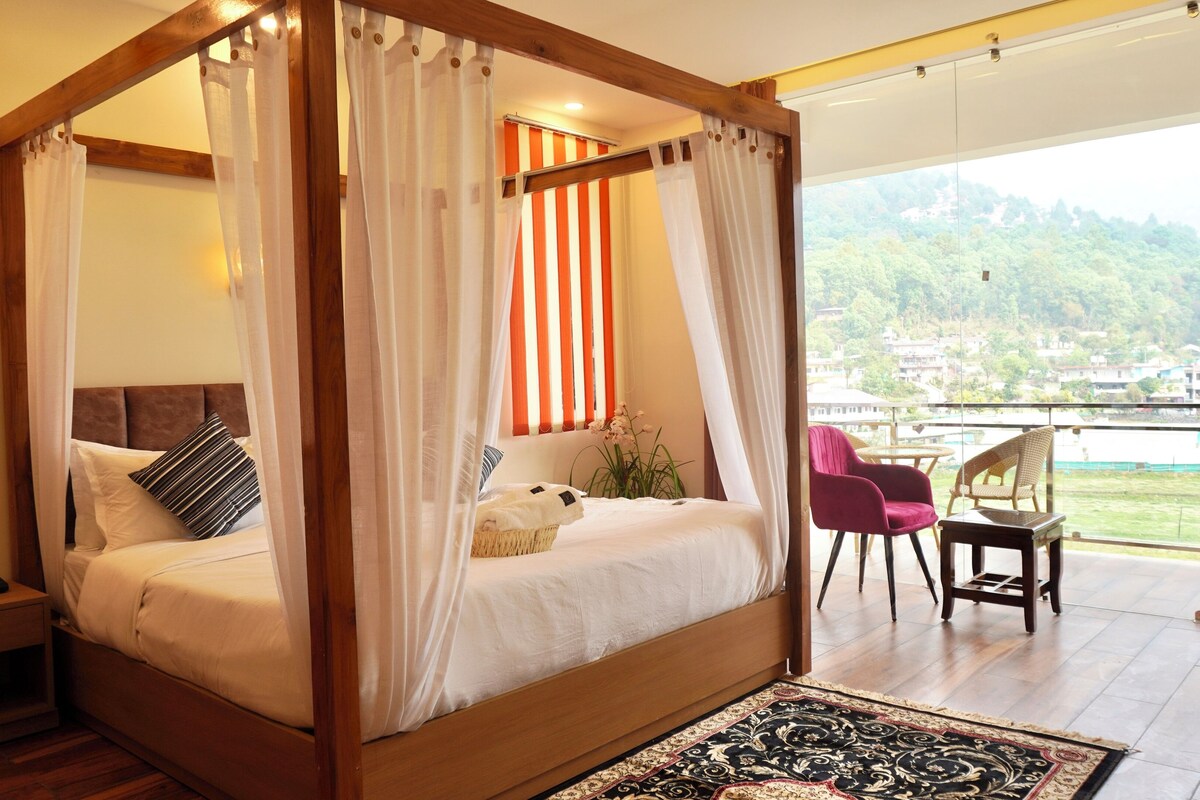 A four-poster bed with sheer curtains, a view of a green landscape, and a balcony with wicker chairs.
