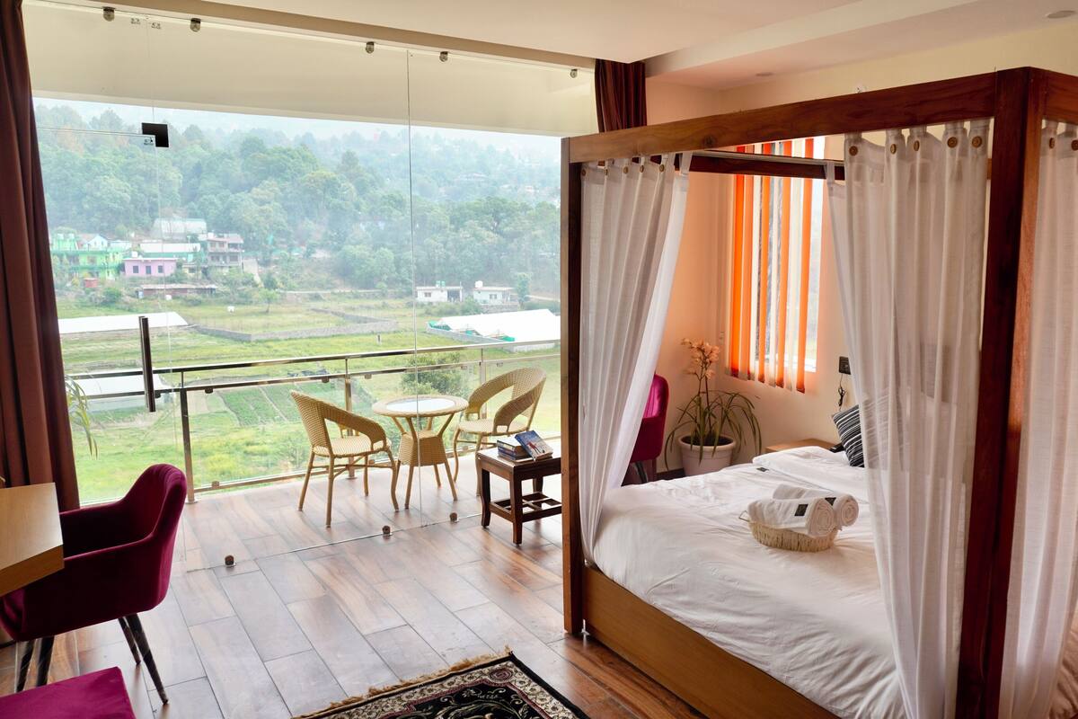 A bedroom with a four-poster bed, a balcony with wicker chairs, and a view of a green landscape.