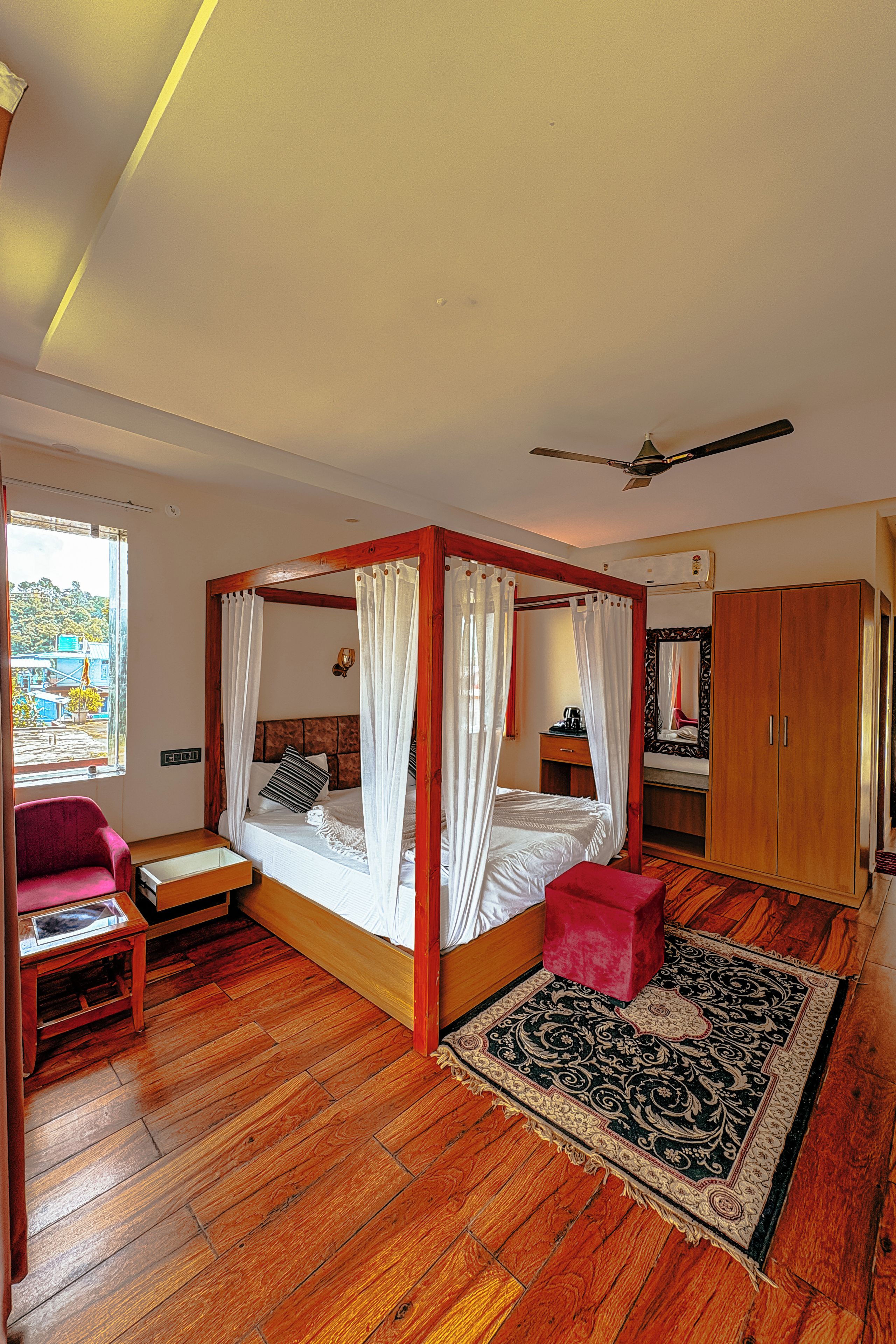 A room with a four-poster bed, a red ottoman, a wooden floor, and a large window with a view.