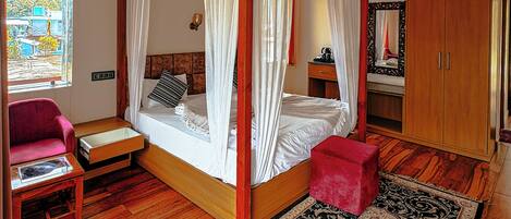 A room with a four-poster bed, a red ottoman, a wooden floor, and a large window with a view.