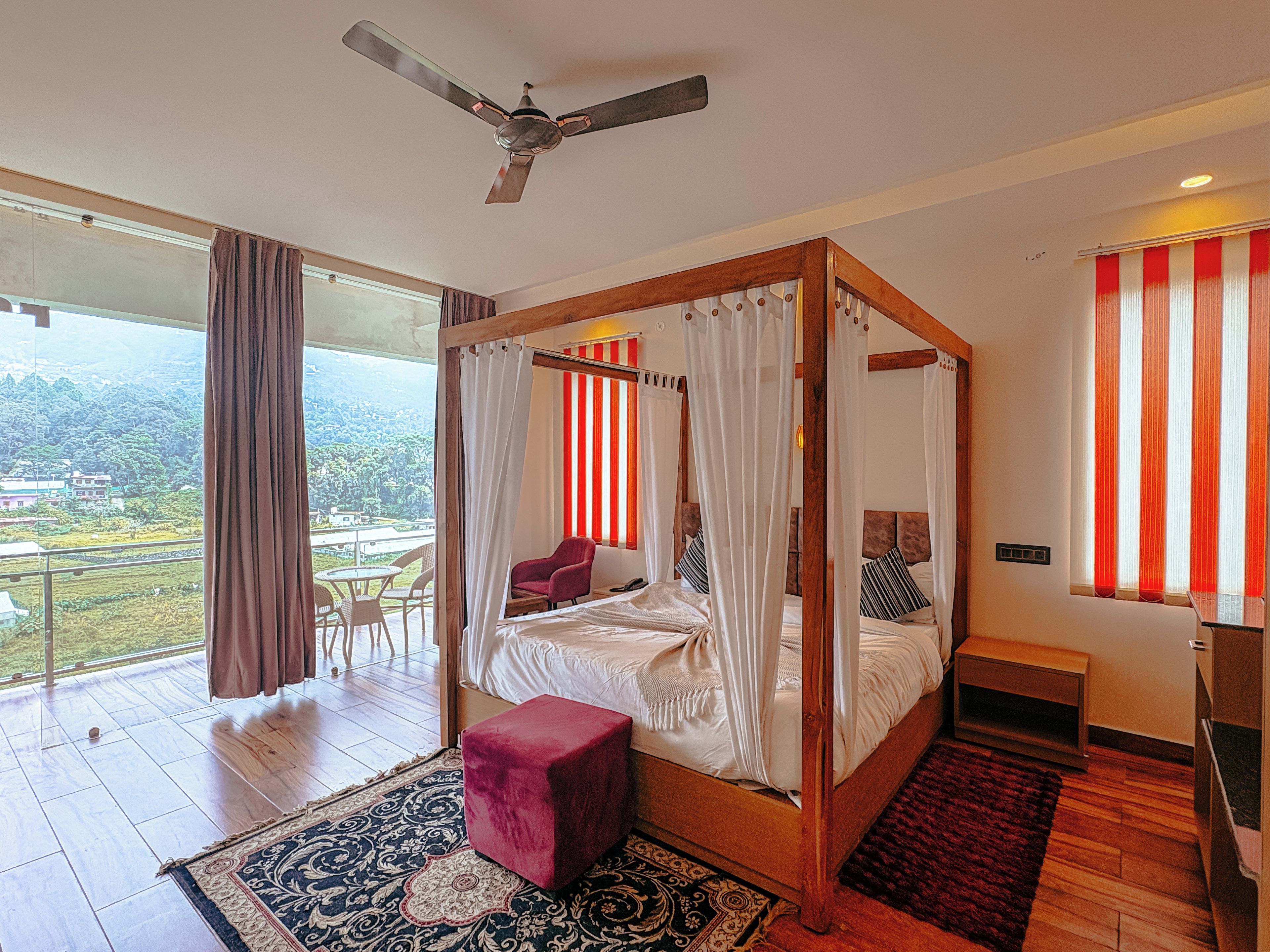 A four-poster bed with a canopy, a red chair, a small wooden nightstand, and a view of a lush landscape through large windows.