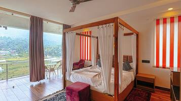 A four-poster bed with a canopy, a red chair, a small wooden nightstand, and a view of a lush landscape through large windows.