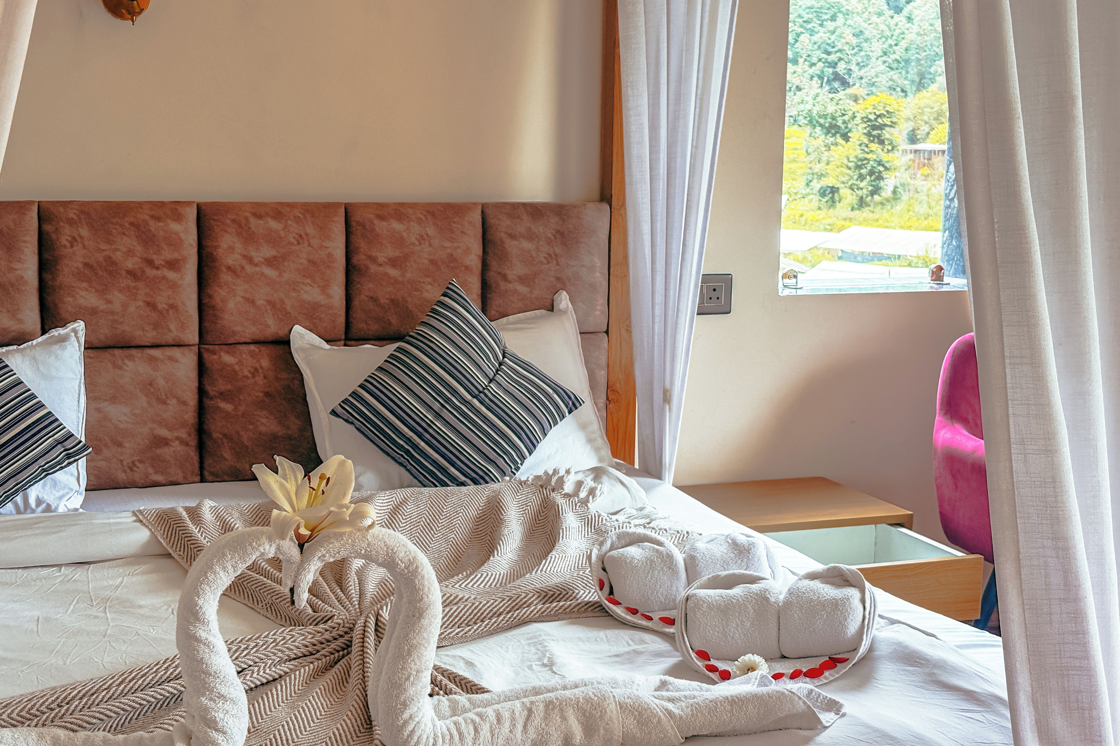 Una habitación de hotel con una cama, almohadas, una mesita de noche, una silla y una ventana con cortinas.